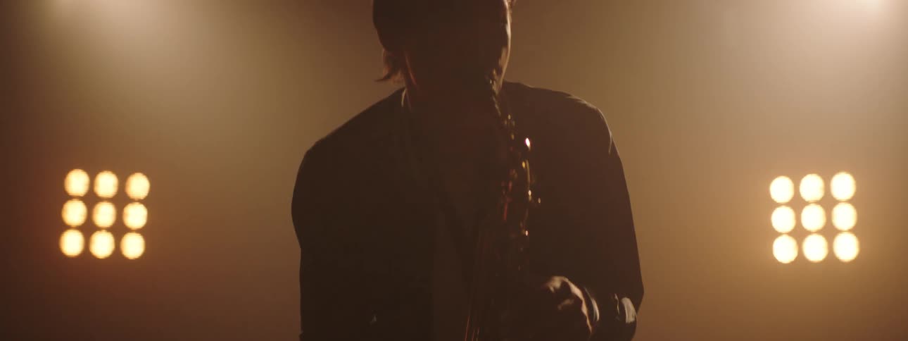 A musician sways as he plays the sax with stage lights glowing behind him