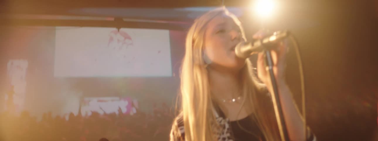 A girl sings with her band in front of a screen showing video of another concert and audience
