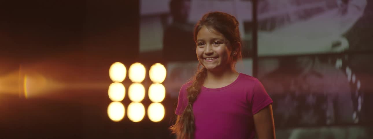 A young girl stands on a stage with a big smile and bright lights