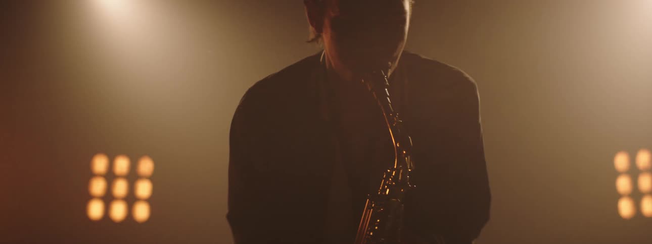 A man sways while playing the saxophone with stage lights shining behind him