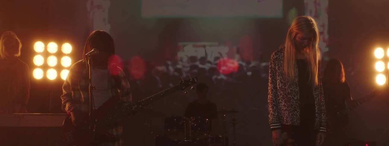 Band members stand still with heads bowed while stage lights glow and video plays behind them