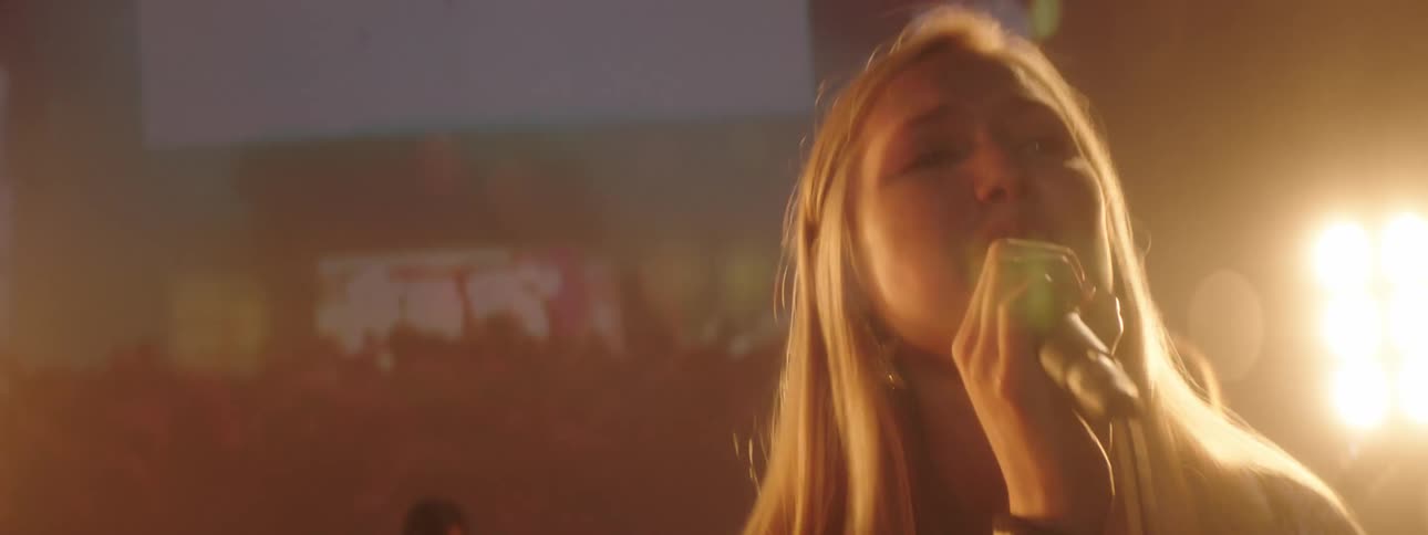 Blonde woman singing on stage with a rock band in front of flashing yellow lights