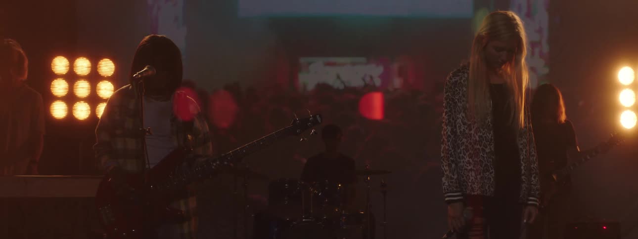 People with instruments standing on a stage with spotlights a screen and a wall in the background