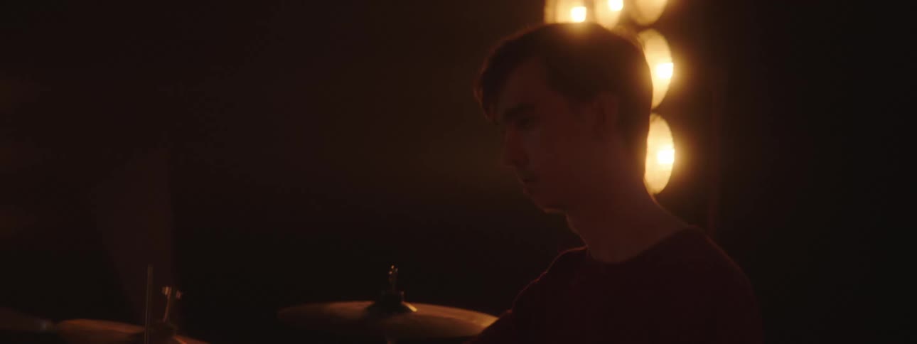 A man in a room playing drums with drumsticks with a wall and spotlights in the background