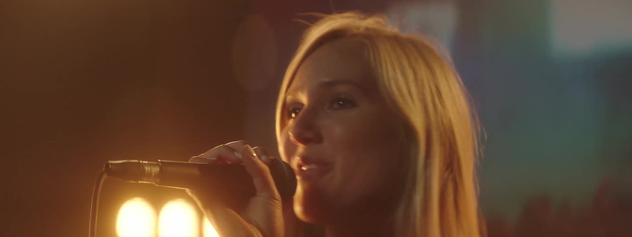 A woman with a microphone singing on stage with spotlights a screen and a wall in the background