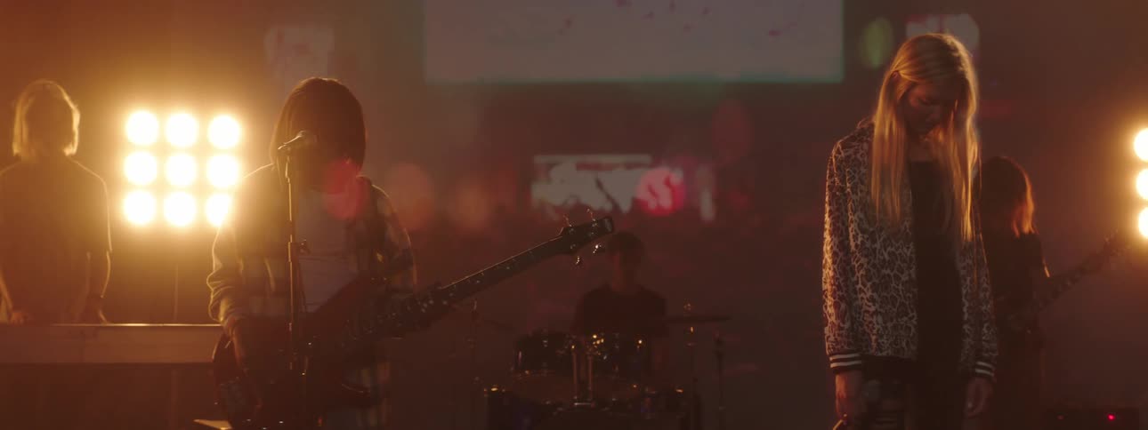 People with instruments standing on a stage with spotlights a screen and a wall in the background