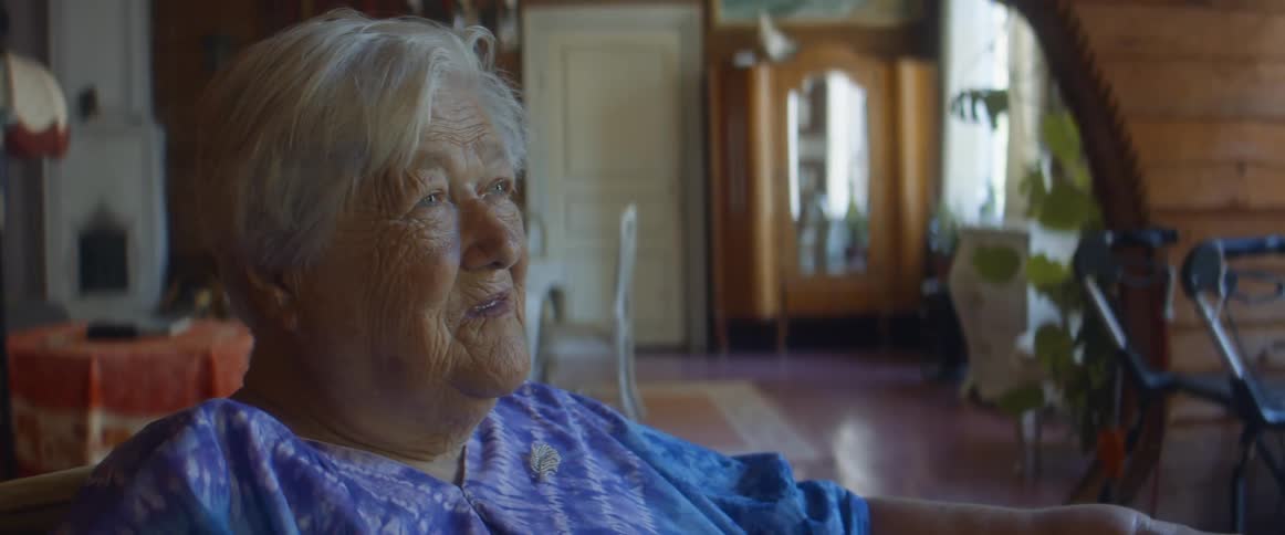 Elderly woman with grey hair blue eyes and blue dress sits in armchair in the living room at home