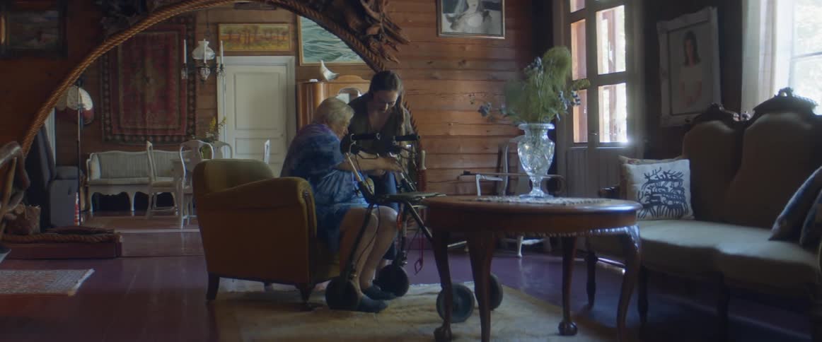 A woman helps her mom get out of an armchair and to her walker