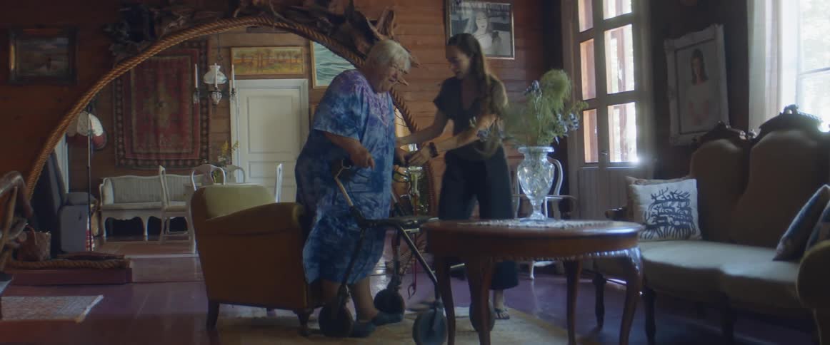 A woman in a room helping an elderly woman walk with a walker with a wall and doorway in the background
