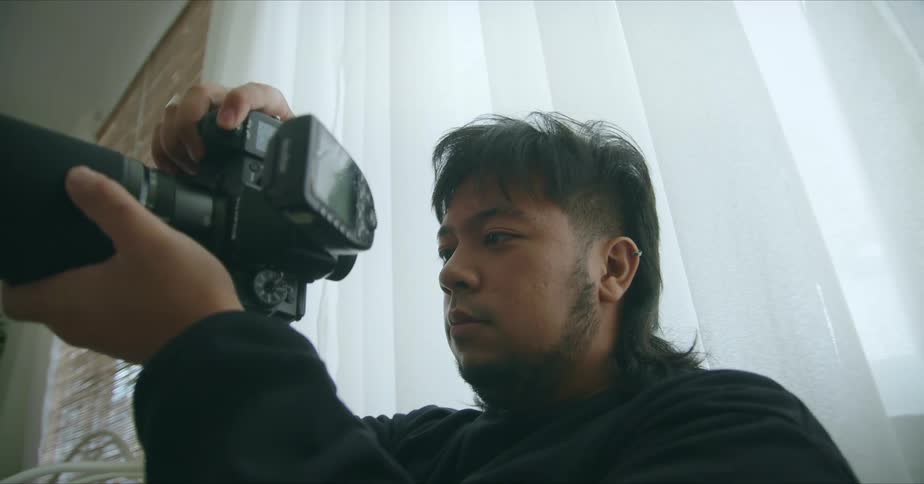 A man holds a film camera with a curtained window behind him