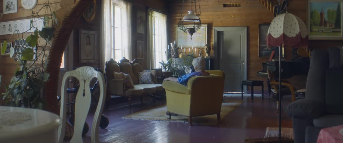 Elderly woman with grey hair and blue dress sits in brown armchair pondering