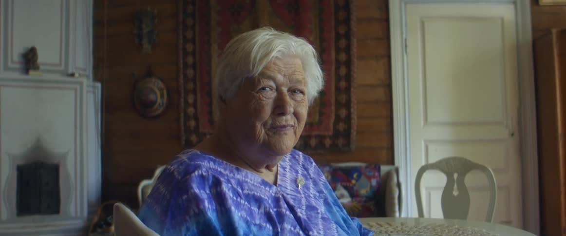 Elderly woman in a blue dress sits in a chair at a table indoors