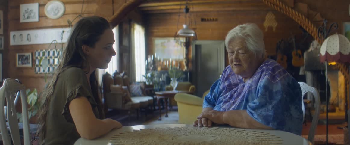A woman points to the crocheted cloth on a table and listens as an elderly woman talks