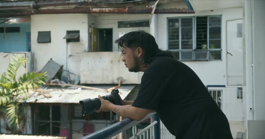 Male photographer with long hair beard and black t shirt leans on railing holding camera