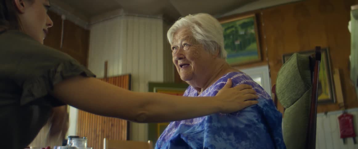 An elderly woman sits in her home chair talking with a young woman beside her