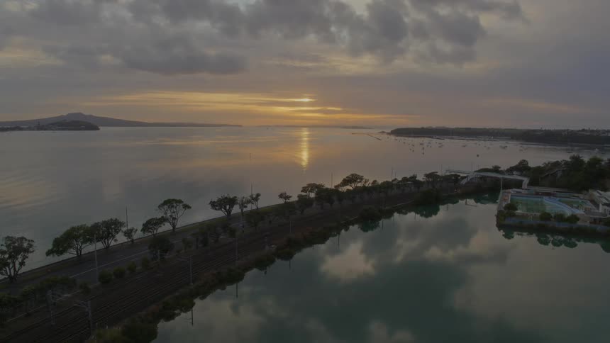 Still water reflects clouds and and obscured sun as bus drives along road past park