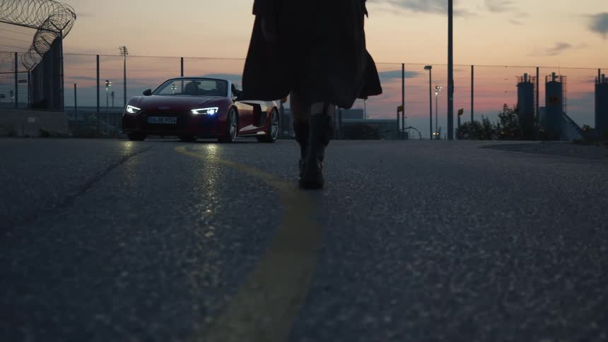 A sports car slows as it approaches a woman walking down the center of a road at sunset
