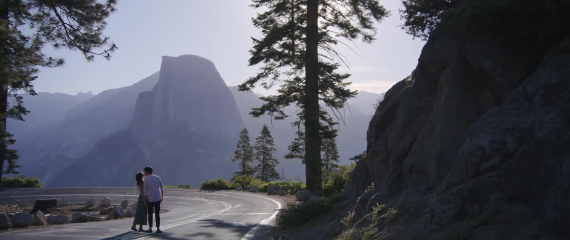 A man and woman stand close together on a mountainside road