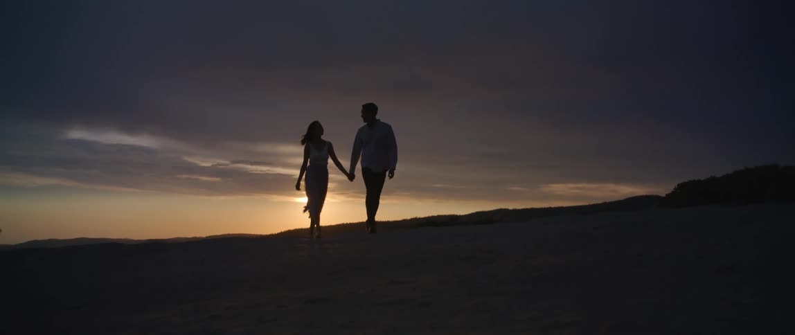 Man and woman hold hands as they walk in a field at dusk