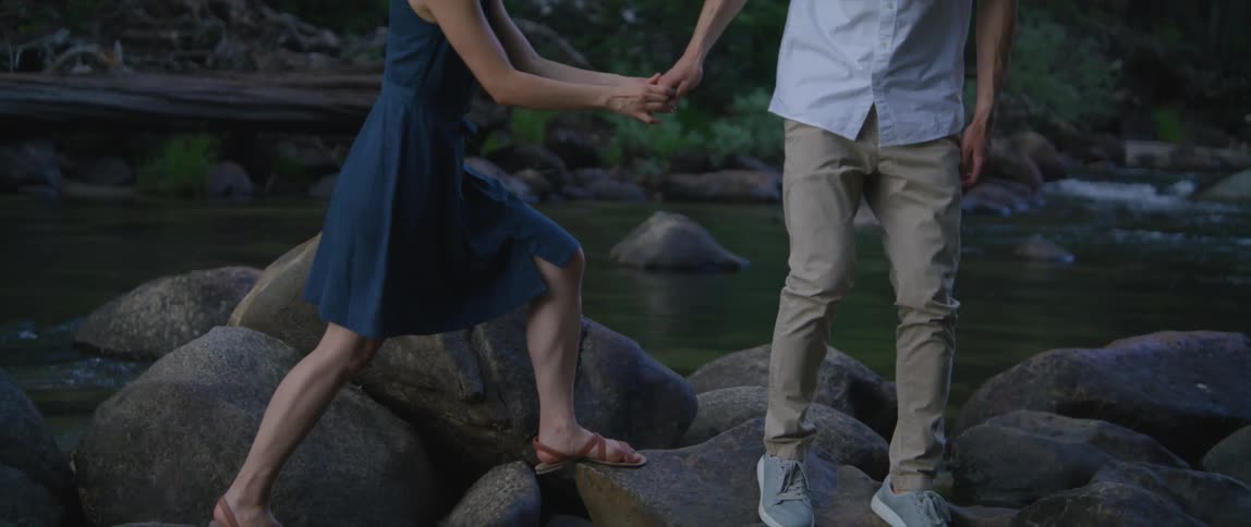 A man and woman are climbing over big rocks near a river