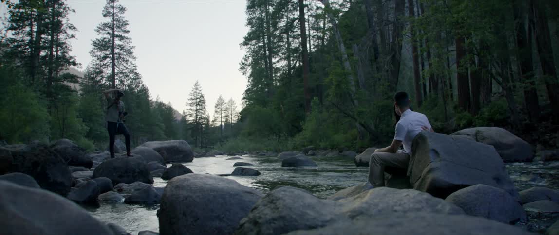 A man and a woman sitting on a rock by river as another man takes a picture with a camera with trees the sky and clouds in the background