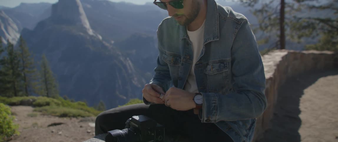 Male photographer with denim jacket black pants and sunglasses sits on wall setting up his camera