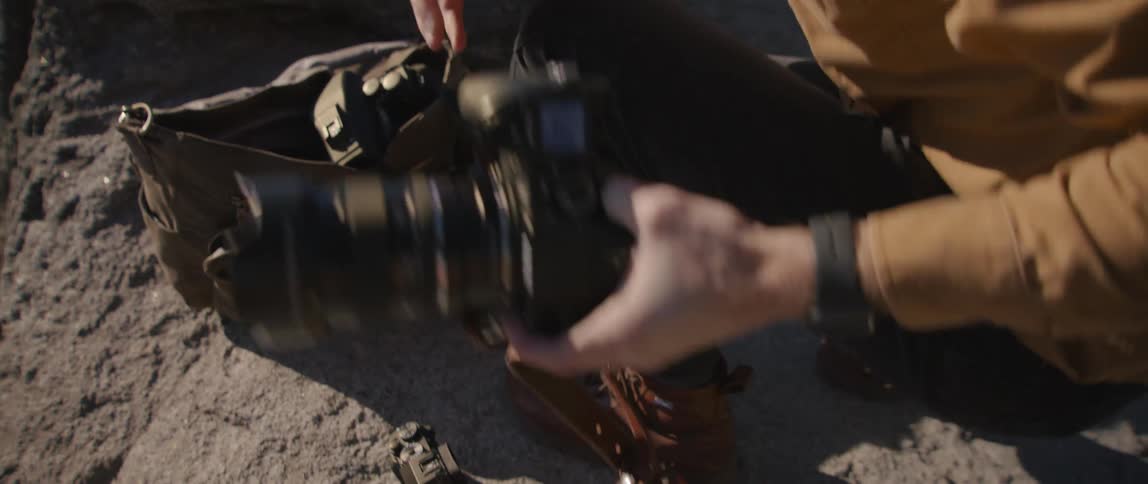 A person on a cliff adjusting a lens on a camera with a mountain range in the background