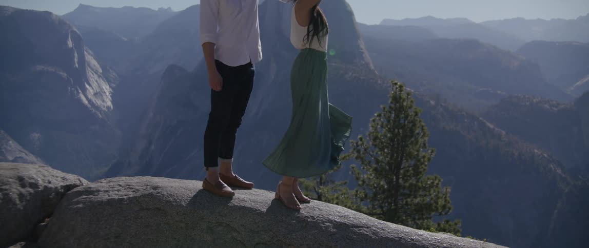 Man spins woman under his hand as they stand on stone overlooking misty valley leading to mountain range