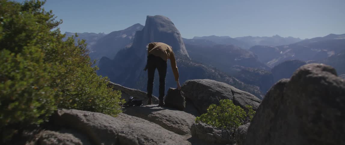 Man with brown shirt and pants carries camera and puts down bag on rock with mountains in the background