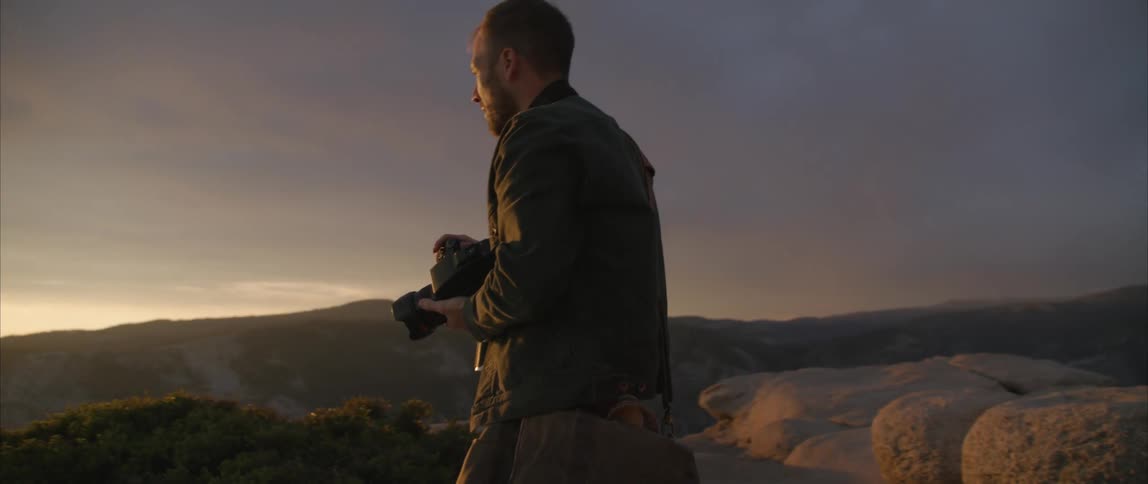 Man with a camera is taking pictures of a couple with mountains and a sunset on the horizon
