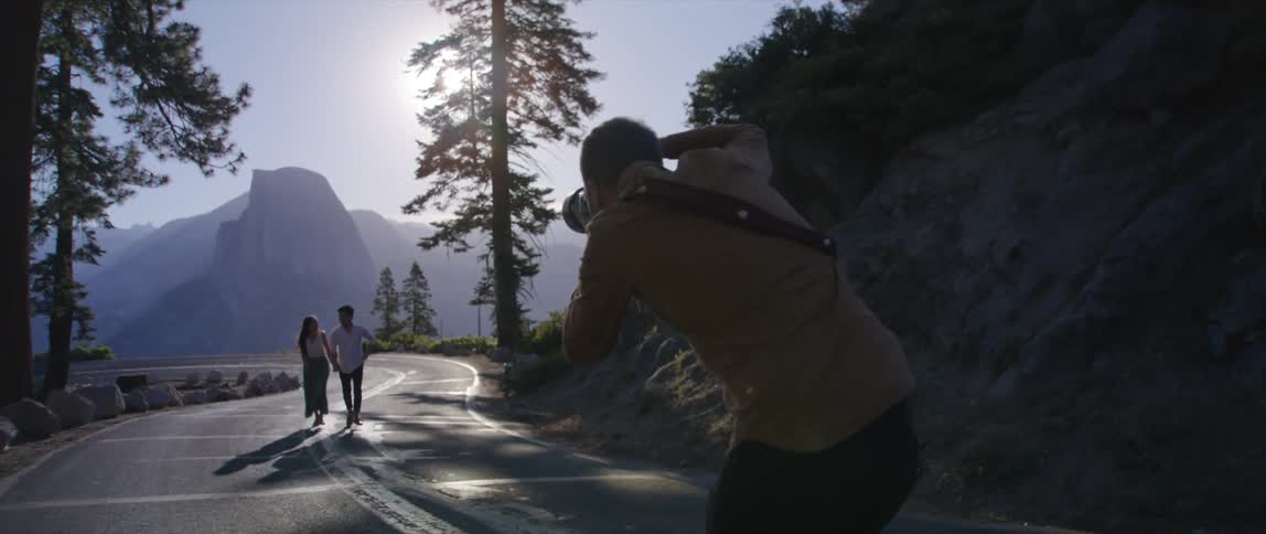 Photographer takes pictures of couple running hand in hand along road with misty mountains in background on sunny day