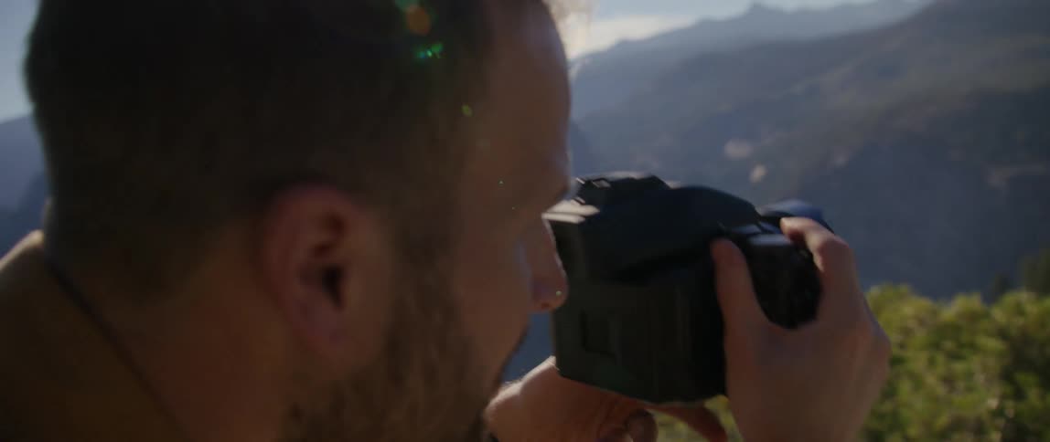 A man with a camera sitting on a cliff taking a picture with a mountain range the sky and clouds in the background
