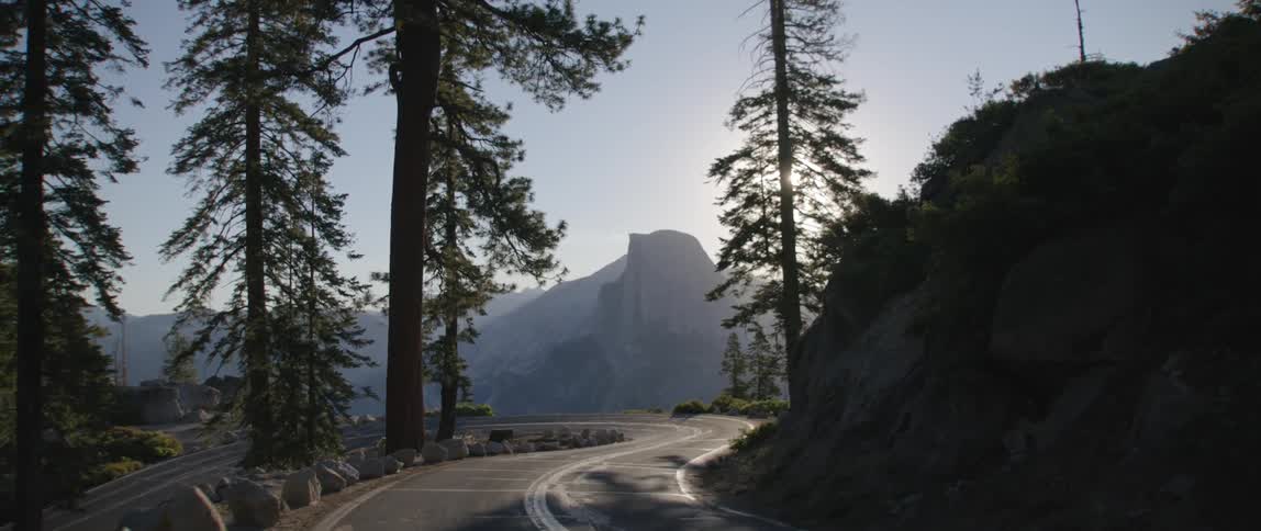 A road with trees by a cliff with a mountain range the sky and clouds in the background
