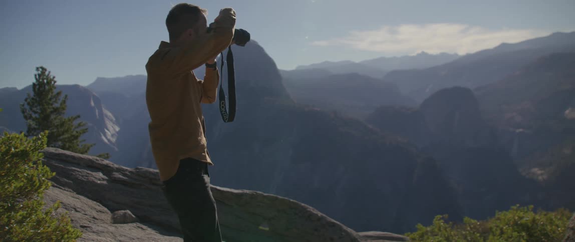 A man crouching along the edge of a huge valley raises his camera to take a photograph of the landscape