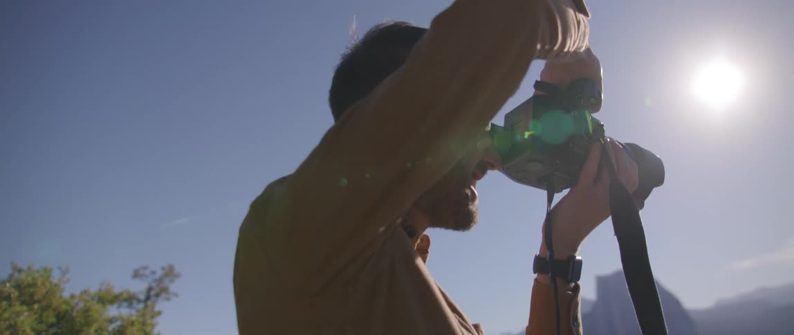 Male photographer holds camera to his eye standing outdoors with mountains in background on sunny day