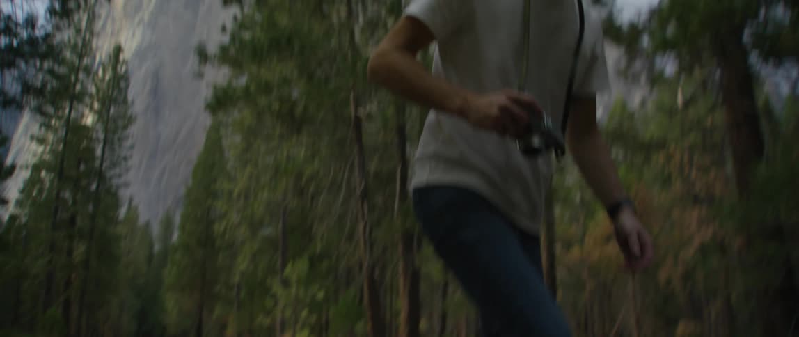 Man wearing dark sunglasses and holding a camera is riding a skateboard down the middle of a forest road with a mountain in the distance