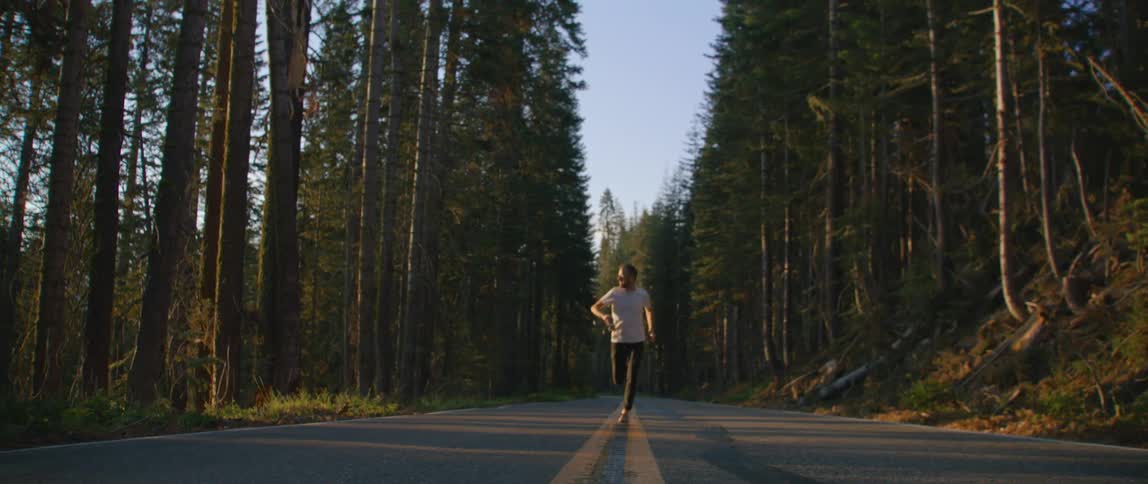 A man with a camera running on a road surrounded by trees with the sky and clouds in the background