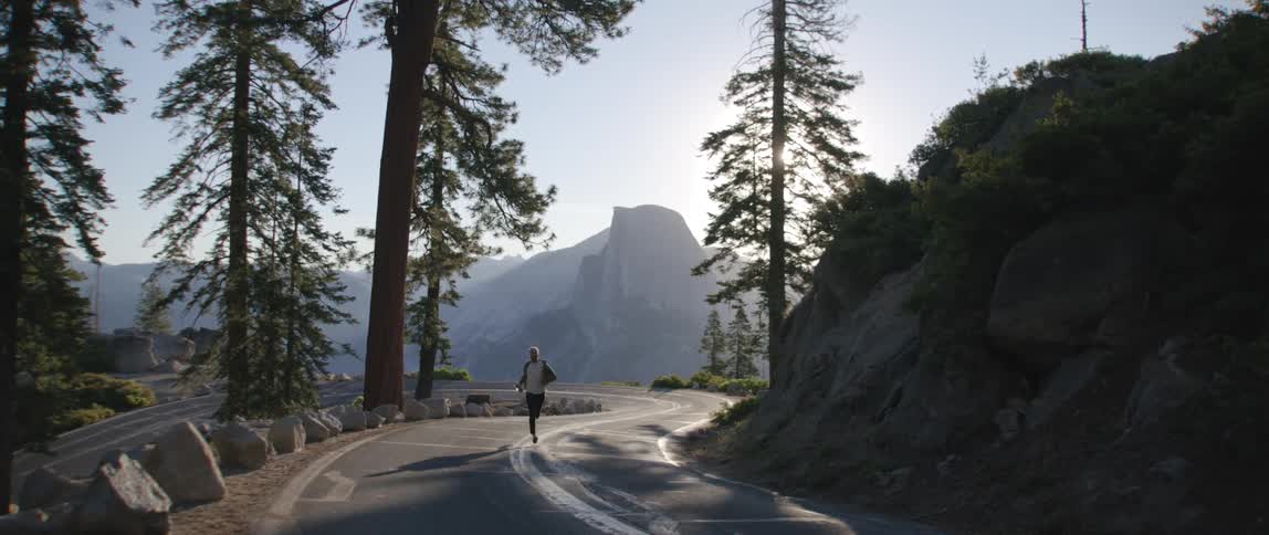 Man runs along empty winding road past slope and tall conifer trees with misty mountain in background
