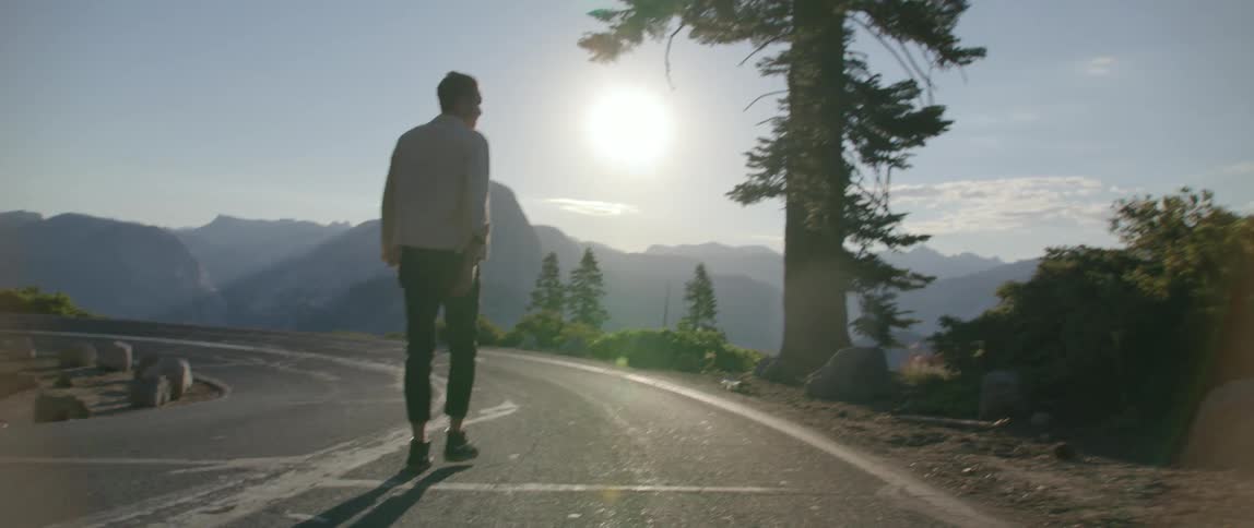 Man walks backward along highway looking toward misty mountain range partly silhouetted by bright sun