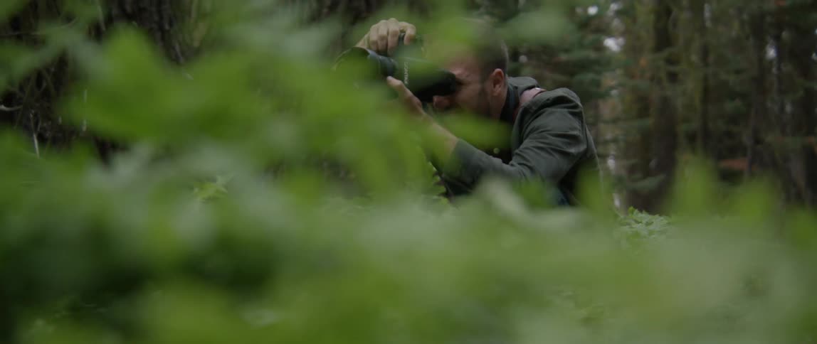 A man in a forest with a camera taking pictures with trees the sky and clouds in the background