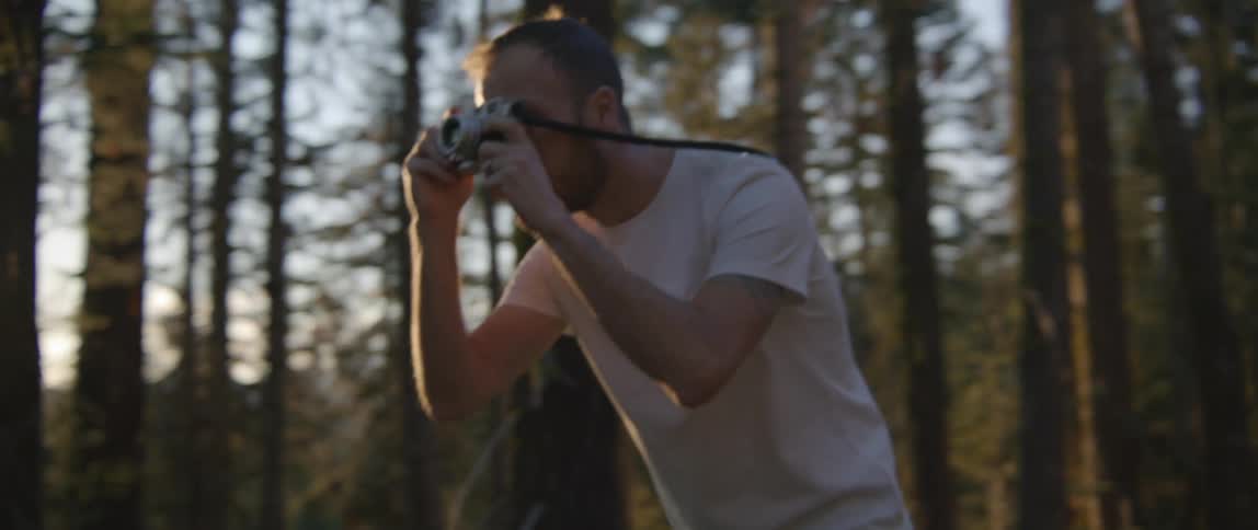 A man with a camera is taking pictures in a forest area with the setting sun shining through the trees