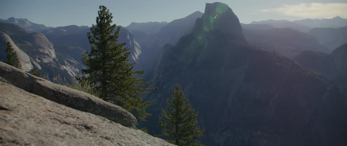 Conifer trees cling to side of steep rocky slope overlooking mountain range stretching to horizon under cloudy sky
