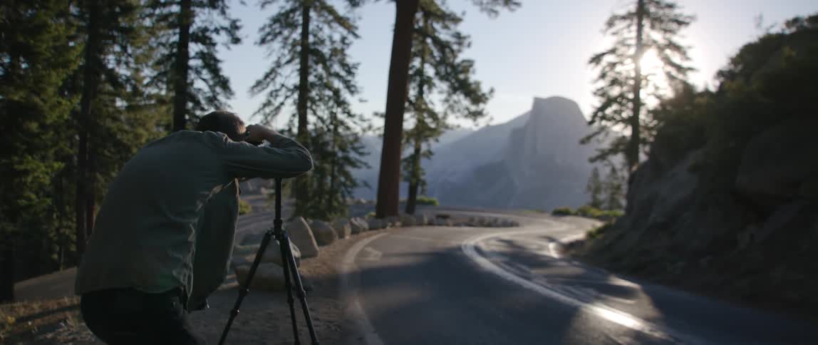 A man with a camera and tripod taking a picture of a road with a mountain range the sky and clouds in the background