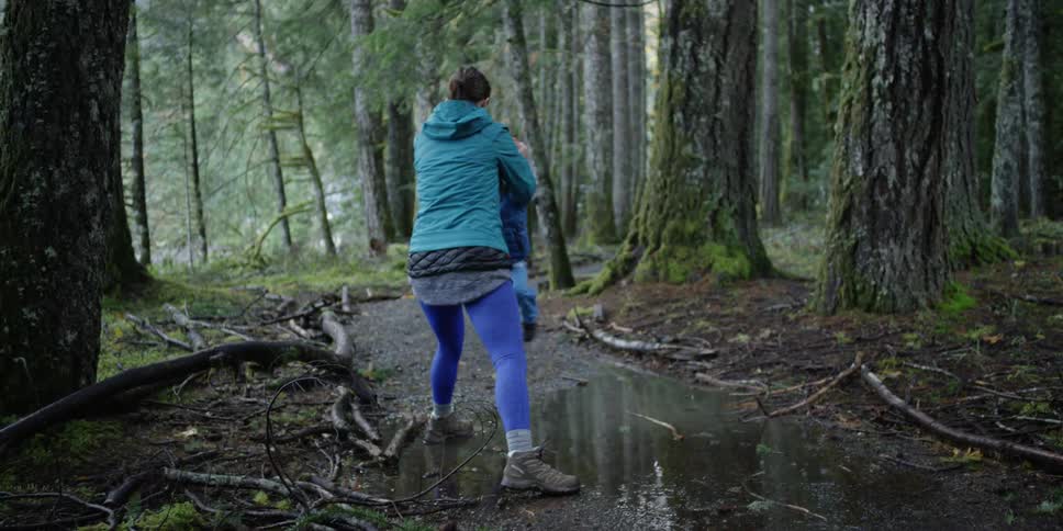 A woman lifts and swings a young boy over a puddle in the woods