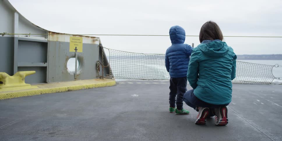 A woman crouches by a child looking at the water from a moving ship