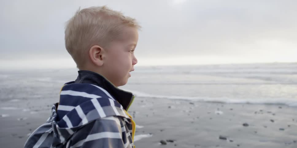 A young boy points to something in the sand as he stands on a beach