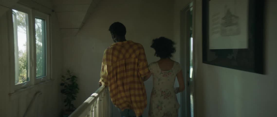 A man and woman hold hands as they run through the narrow corridor of a house towards a staircase