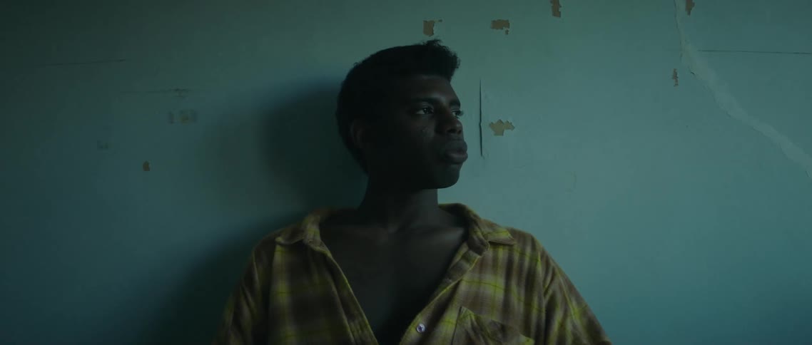 A young man sits on a bed while staring out a window with a thoughtful expression on his face