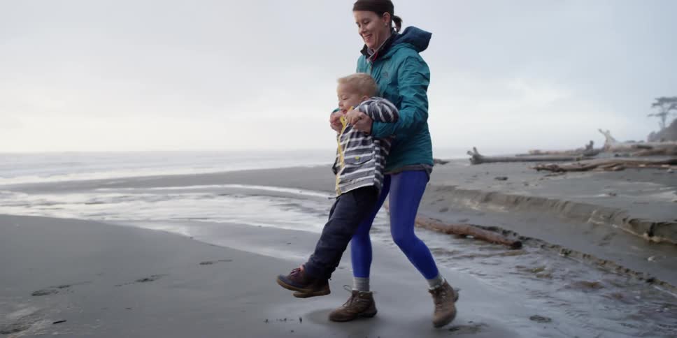 A woman lifts a child over water to walk on a beach