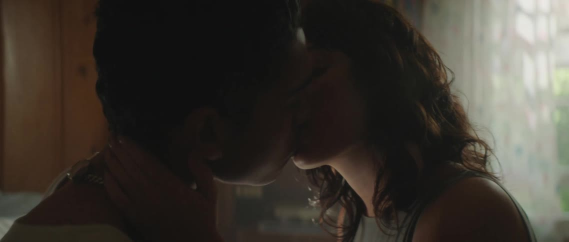 A teenage boy and girl sit on a bed together and kiss one another